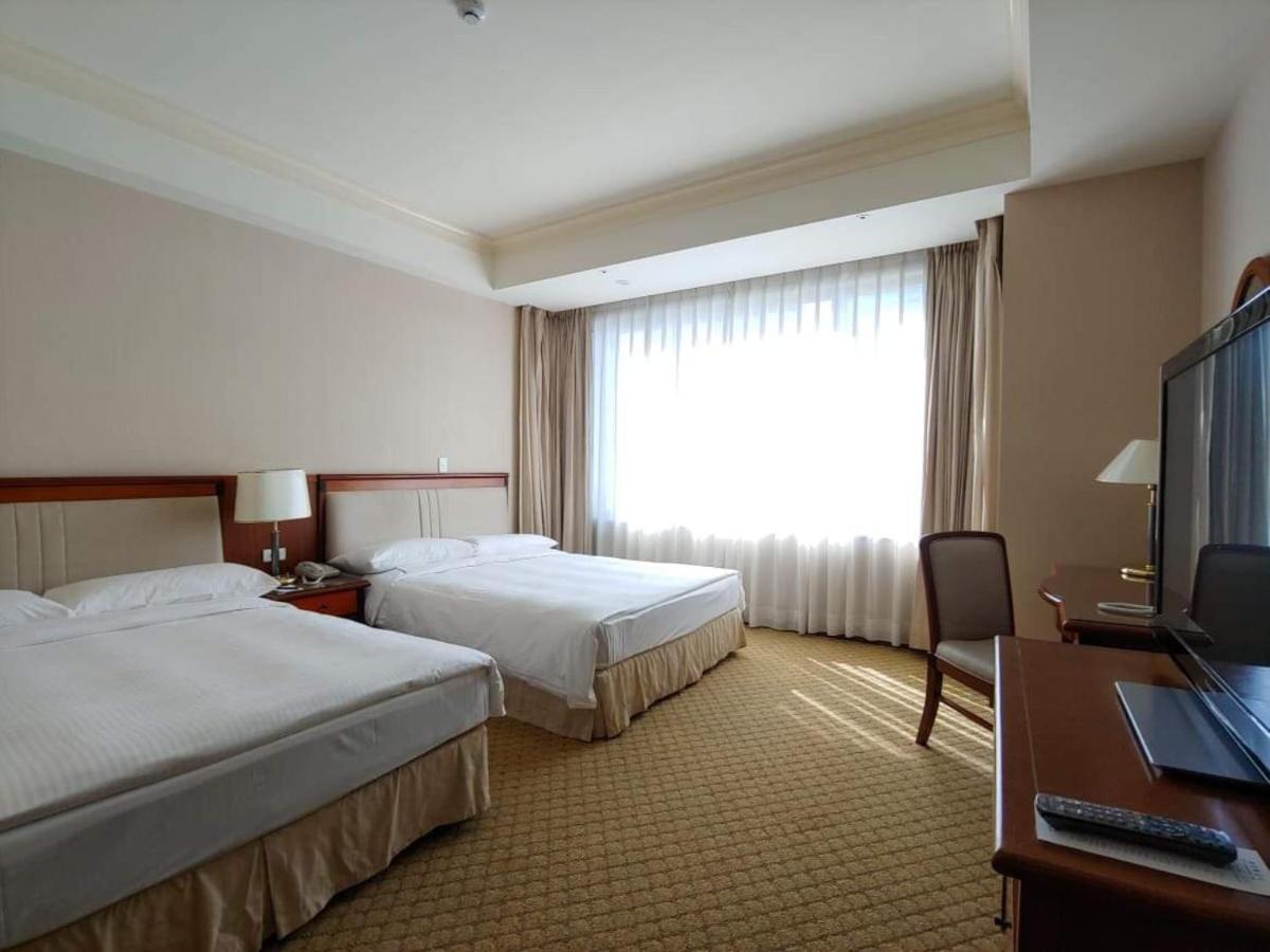 Evergreen Laurel Hotel - טאיצ'ונג מראה חיצוני תמונה The image shows a hotel room featuring two single beds with white bedding. There is a large window with sheer curtains letting in natural light, and the walls are painted in neutral tones. A wooden desk with a chair is positioned near the window, and