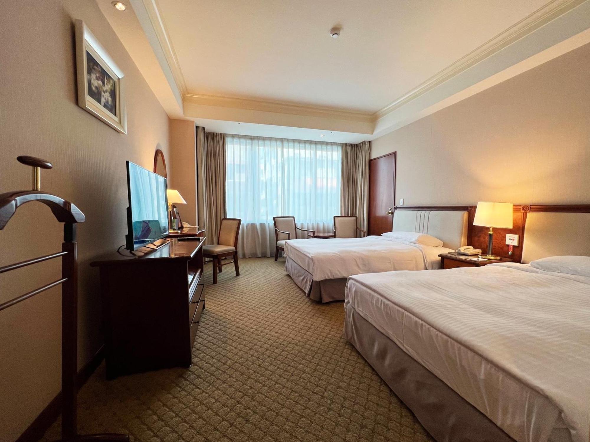 Evergreen Laurel Hotel - טאיצ'ונג מראה חיצוני תמונה The photo shows a hotel room featuring two twin beds placed parallel to each other. The room has a warm color palette and includes a large window with sheer curtains allowing natural light to illuminate the space. There is a wooden desk and two chair