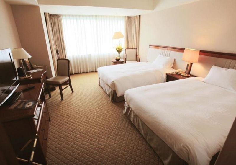 Evergreen Laurel Hotel - טאיצ'ונג מראה חיצוני תמונה The photo shows a hotel room featuring two twin beds with white bedding. There's a chair positioned near one of the beds and a desk with a computer setup against one wall. Large windows allow natural light to enter the room, and light-colored curtain