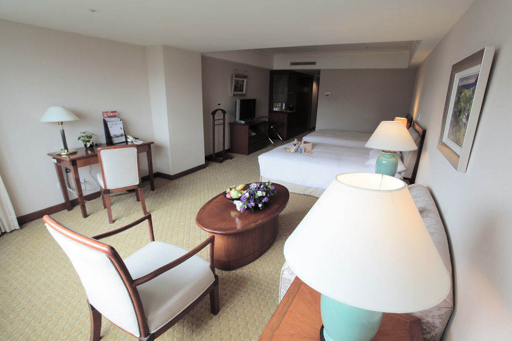 Evergreen Laurel Hotel - טאיצ'ונג מראה חיצוני תמונה The photo shows an interior view of a hotel room. It features two beds, a coffee table with a small decorative arrangement of flowers, and a comfortable chair. There's a desk and a lamp, along with another larger lamp on the table. In the background,