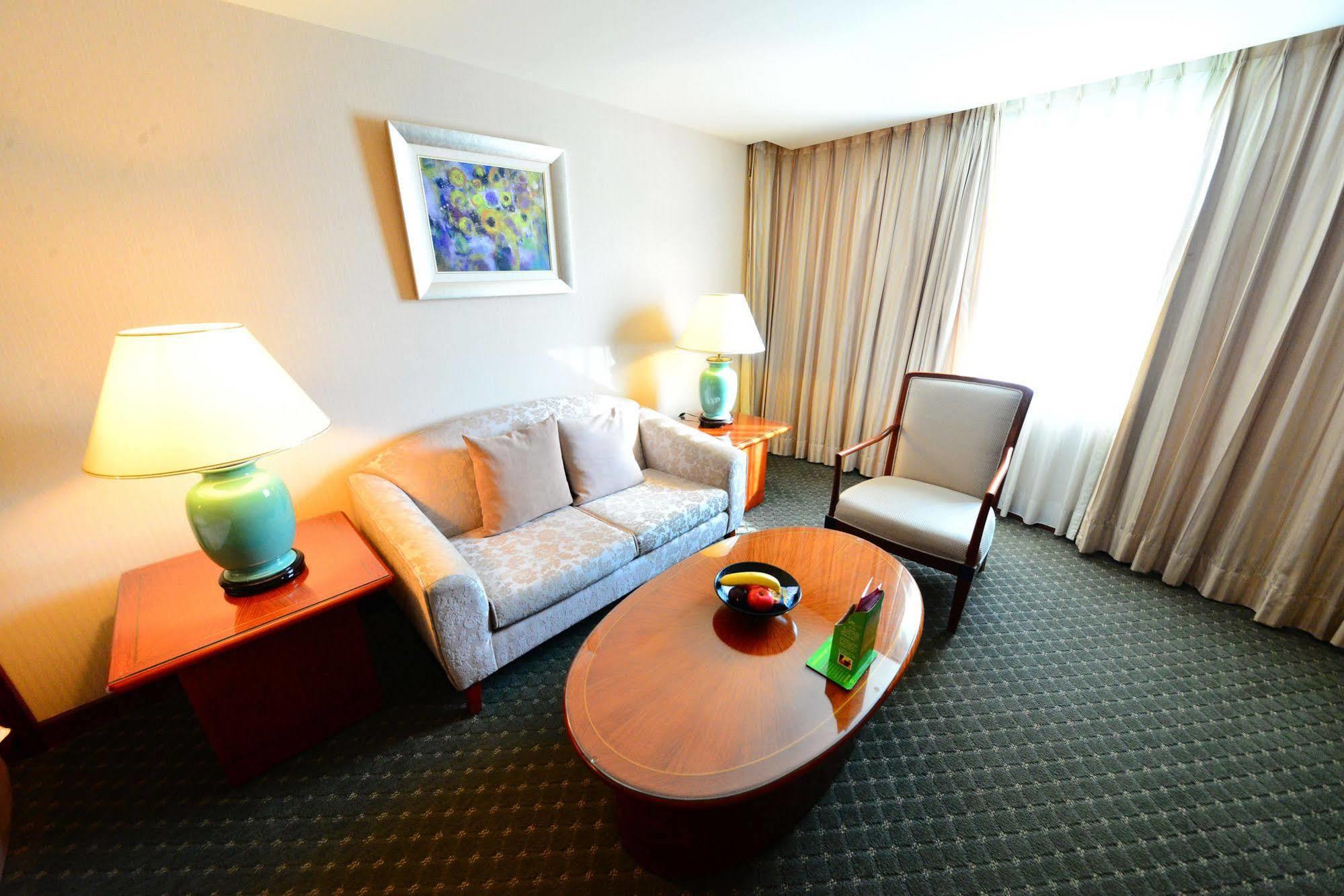 Evergreen Laurel Hotel - טאיצ'ונג מראה חיצוני תמונה The photo shows a cozy living space, likely a hotel room or a lounge area. It features a light-colored sofa with cushions, a wooden coffee table in the center, and a comfortable armchair beside it. There are two lamps with turquoise bases on side tab