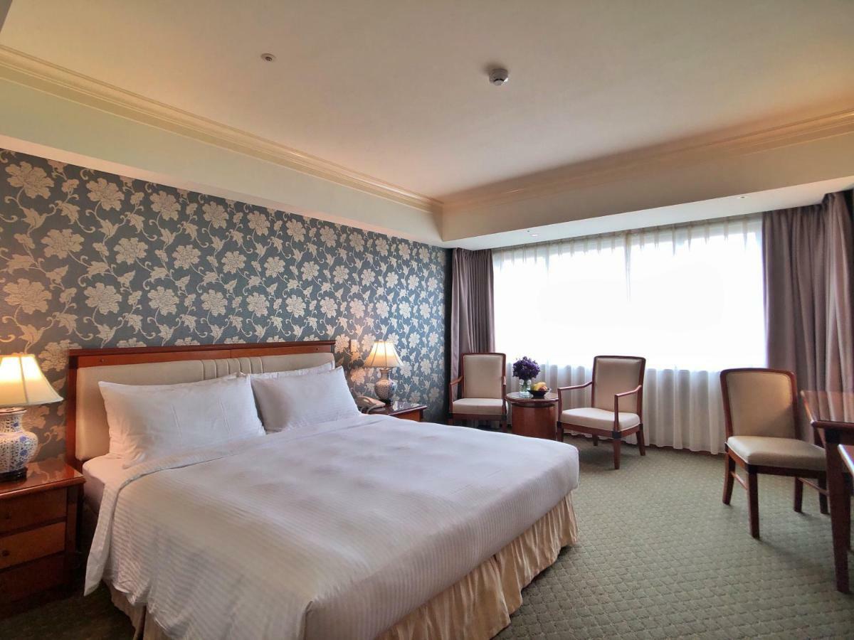 Evergreen Laurel Hotel - טאיצ'ונג מראה חיצוני תמונה The photo shows a hotel room that features a large bed with a white bedspread positioned against a decorative floral wallpaper. There are two bedside lamps on wooden nightstands flanking the bed. To the side of the room, there are two chairs and a sm