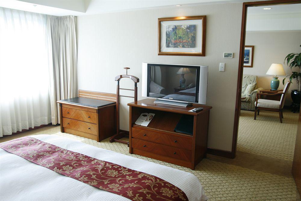 Evergreen Laurel Hotel - טאיצ'ונג מראה חיצוני תמונה The photo depicts a hotel room interior. In the foreground, there is a neatly made bed with a decorative red bedspread. To the left, a large window allows natural light to fill the room, with light-colored curtains drawn to one side. 

Next to the be